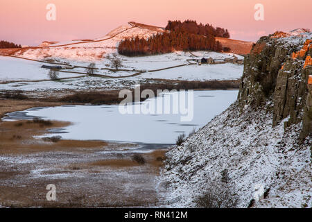 Winter Schnee liegt auf den Feldern und Felsen neben dem gefrorenen Crag Lough auf dem Hadrianswall in Northumberland. Stockfoto