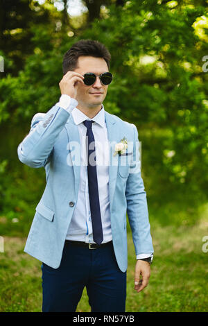 Schönes Modell Mann, in der modischen blauen Jacke mit einer schmalen Krawatte und stilvolle Sonnenbrillen im grünen Garten steht Stockfoto