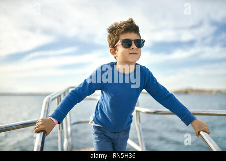 Freundliche kleine Junge in Sonnenbrille auf Yacht posiert an sonnigen Meer Hintergrund Stockfoto