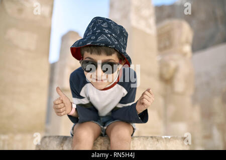 Fratzenhaft kleiner Junge in Sonnenbrille sitzt auf den Stein auf die unscharfen sonnigen Hintergrund der antiken Ruinen in Luxor in Ägypten. Er trägt ein blaues - Red Hat wi Stockfoto