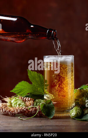 Bier ist in einem Becher aus der Flasche gegossen. Becher Bier, Korn und Hopfen auf einem braunen Hintergrund. Freier Platz für Text. Stockfoto