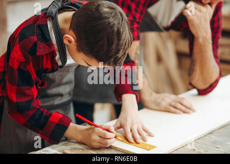 Fleißige kleine Junge besetzt ist, Messen und Markieren von Holzbrett mit einem Bleistift mit seinem Vater als Zimmermann sitzend in Holz- worksho Stockfoto