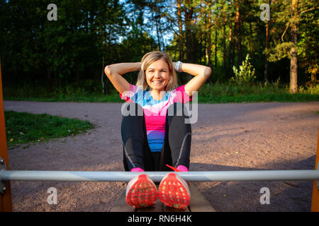 Athlet Blondine mit den Händen hinter dem Kopf üben auf Holzbank im Park im Sommer.