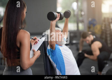 Professionelle personal trainer arbeiten mit Klienten in der Turnhalle, Beobachten mit Ordner und Stift, schreiben. Älterer Mann mit grauen Haaren sitzen, halten sich an den Händen und Übungen mit Hanteln. Stockfoto