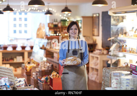 Portrait von Lächelnd weibliche Besitzer von Feinkost Shop tragen Schürze Holding Laib Brot Stockfoto