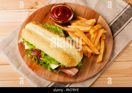 Frische submarine Sandwich mit Schinken, Käse, Speck, Tomaten und Salat auf Holz Schneidebrett mit Pommes frites und Sauce, Ansicht von oben Stockfoto