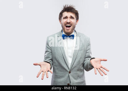 Warum? Was? Portrait von verwirrt aussehenden bärtigen Mann in lässigen grauen Anzug, blau Bow Tie stehen und von der Kamera mit erhobenen Armen und Fragen. ind Stockfoto
