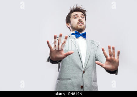 Portrait von schockiert aussehenden bärtigen Mann in lässigen grauen Anzug und blau Bow Tie stehend mit Blockierung Hände und Suchen bei Camera erschrocken. indoor Studio Stockfoto