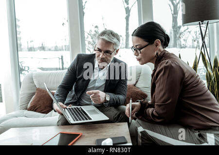 Hohe grauhaarige gut aussehender Mann in einer dunklen Jacke und seine Partner suchen ernsthafte Stockfoto