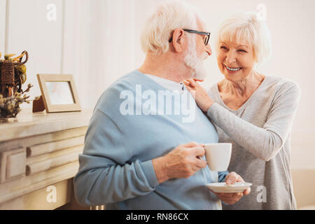 Liebevolle Umarmung. Ältere lächelnde Frau Gefühl unvergesslich, während Ihr liebevoller Mann, der leckere Kaffee umarmen Stockfoto