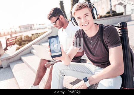 Mein Gadget. Attraktive junge halten Lächeln auf seinem Gesicht und halten Telefon während auf Kamera posieren Stockfoto