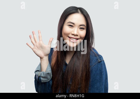 Portrait von glücklich schöne Brünette asiatische junge Frau in Blau Jeansjacke, Make-up-winkenden Hand und Gruß an die Pflege von Kamera schaut. Stockfoto