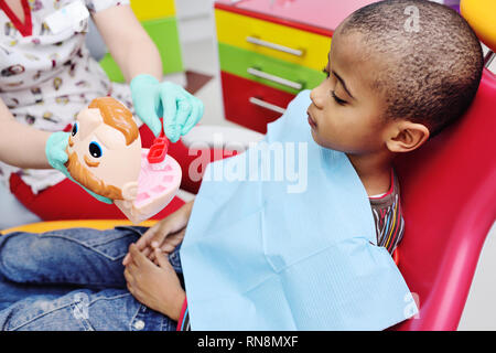 Ein pädiatrischer Zahnarzt lehrt einen afrikanischen amerikanischen Kind, das in einem Behandlungsstuhl sitzt, um seine Zähne zu putzen. Stockfoto