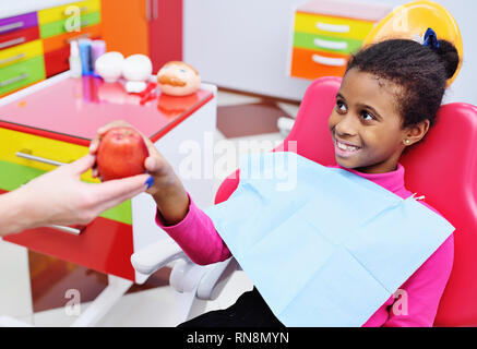 Zahnarzt gibt eine reife rote Apple zu einem kleinen schwarzen afrikanischen amerikanischen Mädchen in einem behandlungsstuhl. Stockfoto