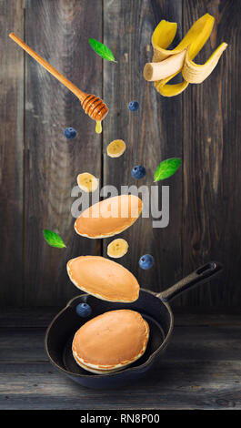 Hausgemachte Pfannkuchen, Blaubeeren und Banane in eiserne Pfanne fliegen mit rustikalen Holzmöbeln Hintergrund. Stockfoto