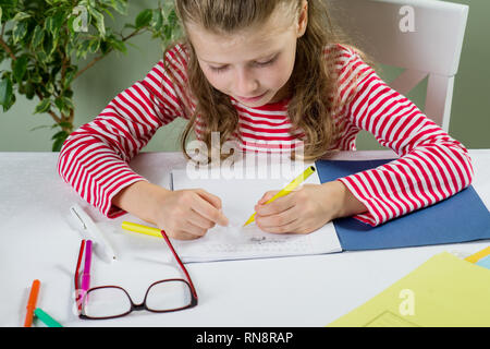 Ein jüngeres Schulmädchen mit Brille schreibt etwas mit der linken Hand auf dem Notebook- und setzt sich an den Tisch. Zurück zum Konzept der Schule. Das Kind ist links Stockfoto