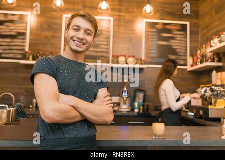 Junge männliche Barista Coffee shop Arbeitnehmer lächelnd in die Kamera schaut mit verschränkten Armen in der Nähe der Theke gekreuzt. Stockfoto