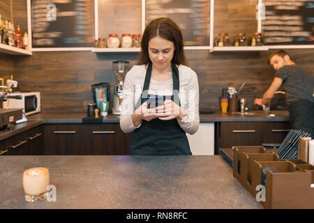 Junge Frau in Schürze coffee shop Arbeiter am Tresen, um auf dem Smartphone. Stockfoto