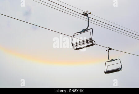 Zwei leere Skilift Stühle auf Kabel bei bewölktem Himmel ausgesetzt, die mit einem gelben und rosa Band von Sonnenlicht Stockfoto