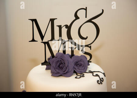 Nahaufnahme der Herr und Frau Hochzeitstorte am Hochzeitstag. Kuchen hat weiße Zuckerglasur, lila Band und Puderzucker lila Blumen es dekorieren. Stockfoto