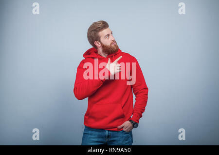Die Jungen gut aussehenden bärtigen Mann mit Rot sweatshirt stehend auf dem grauen Hintergrund seine Hand in die Tasche und zeigen mit dem Finger auf der rechten Seite Stockfoto