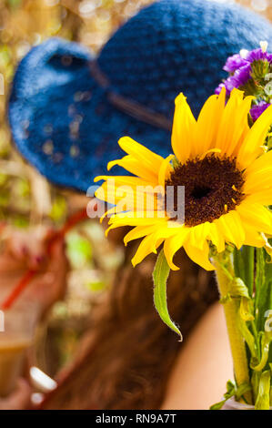 In der Nähe von blühenden lebendige gelb Sonnenblume mit unscharfen Frau trägt blaue Sommer Hut trinken Cocktail mit roten Stroh auf dem Hintergrund Stockfoto