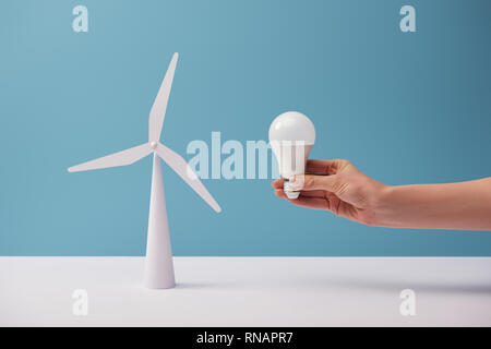 7/8-Ansicht von Frau mit Glühbirne über weiß Tisch mit windmühle Modell auf blauem Hintergrund Stockfoto