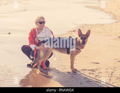 Schönen Witwe ältere Frau mit ihr spielen Deutscher Schäferhund Hund genießen ihre Begleiter und Liebe am Strand bei Sonnenuntergang in Vorteile von Tieren acti Halten Stockfoto