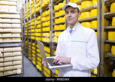 Technologe in einem weißen Mantel mit einem Laptop in der Hand ist im Lager von Käse in der Werkstatt für die Herstellung von Butter und Käse. Qualität con Stockfoto