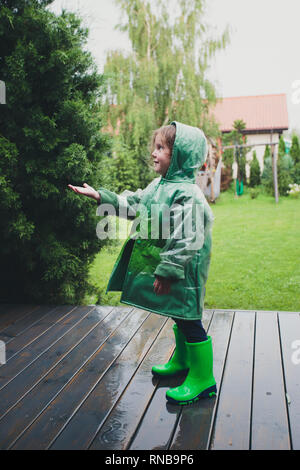 Glücklich lächelnde Mädchen stehen auf einer Veranda fangen Regentropfen tragen grüne Regenmantel Stockfoto