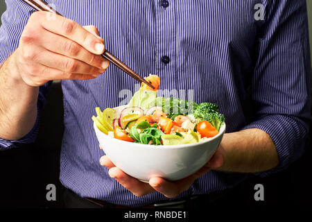 Mann in einem T-Shirt hält poke Schüssel mit Lachs, Avocado, Gurken, Rucola, Brokkoli, Reis, Karotten, und chuha mit Stäbchen Stockfoto