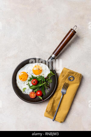 Schwarze Pfanne mit Spiegelei, frische Kräuter und Tomaten, die Schaltgabel und die Serviette auf weißen Hintergrund. Konzept der gesunden Ernährung. Ansicht von oben, freier Speicherplatz Stockfoto