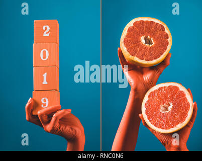 Collage mit Holzwürfeln mit 2019 Nummern und Grapefruit Hälften in Coral farbige weibliche Hände auf Blau getrennt Hintergrund, Farbe 2019 Konzept Stockfoto