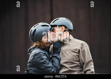 Active Senior Paar mit Fahrrad Helme stehen im Freien againts dunklen Hintergrund, küssen. Stockfoto