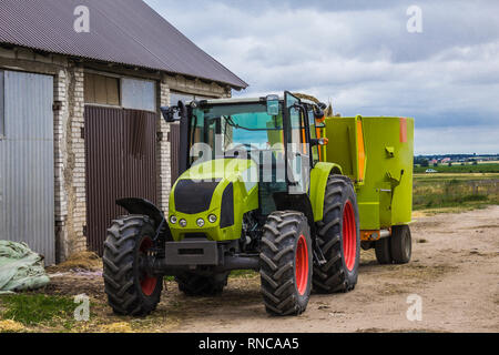 Traktor mit Anhänger für das Mischen und die Verteilung von Futtermitteln für Kühe. Hinter dem Traktor ist eine Scheune und Felder. Notwendige Ausrüstung für eine Molkerei. Stockfoto