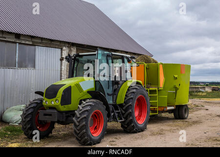 Traktor mit Anhänger für das Mischen und die Verteilung von Futtermitteln für Kühe. Hinter dem Traktor ist eine Scheune und Felder. Notwendige Ausrüstung für eine Molkerei. Stockfoto