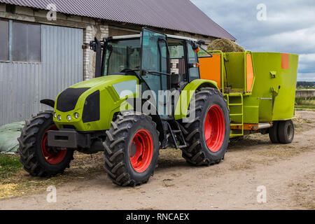 Traktor mit Anhänger für das Mischen und die Verteilung von Futtermitteln für Kühe. Hinter dem Traktor ist eine Scheune und Felder. Notwendige Ausrüstung für eine Molkerei. Stockfoto