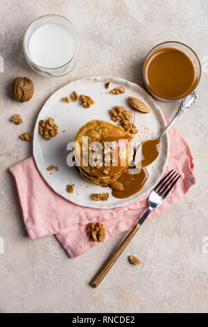 Stapel von gebackenen Pfannkuchen mit gekochten Kondensmilch und Walnüsse, auf weiße Platte mit Gabel, Messer, Mandel, rosa Serviette, jar von Karamell und Milch in Glas Stockfoto