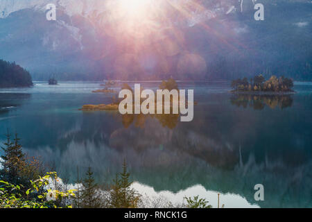 Erste Sonnenstrahlen auf dem Berg Eibsee in den Bayerischen Alpen. Sonnenaufgang über Berge und kleine Insel mit Reflexion, Deutschland, Europa Stockfoto