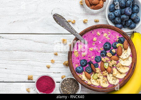 Acai smoothie Schüssel gefüllt mit Banane, Chia und Kürbiskerne, Blaubeere, Mandeln und Müsli mit Löffel auf weißem Holz- Hintergrund. gesund vegan Brea Stockfoto