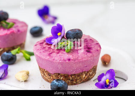 Heidelbeeren und Acai vegan Cashew Kuchen mit frischen Beeren, essbare Blüten, Minze, Muttern. Gesunde vegane Ernährung Konzept. Nach oben Schließen Stockfoto