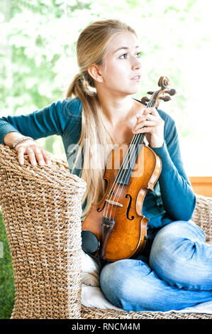 Junge Frau mit langen, blondn Haaren Sitz in einem sitzmoebel aus Ratan und haelt eine Geige auf dem Schoss. [(C) Dirk Friedrich A. Stockfoto