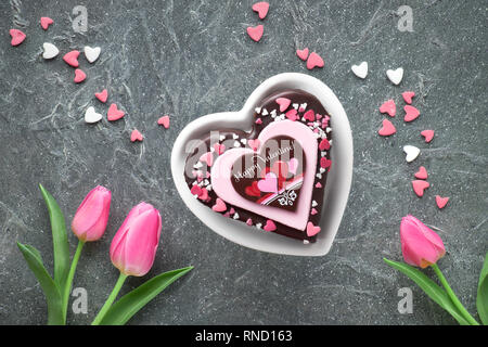 Valentine Herz Kuchen mit Schokolade und Zucker Dekorationen und Gruß Text "Happy Valeitine' und rosa Tulpen. Draufsicht auf dunklem Hintergrund Stockfoto