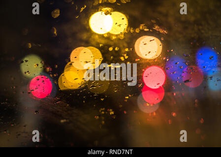 Fokus auf den Vordergrund. Regen fällt auf das Fenster des Autos. Der Verkehr und die Lichter der Stadt im Hintergrund. Stockfoto