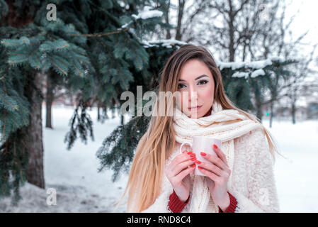 Schöne Mädchen Winter Forest, Hintergrund Fichte und Weihnachtsbaum. Wickelte Strickschal, starker Frost in der Natur. Hand Tasse heißen Kaffee oder Tee Stockfoto