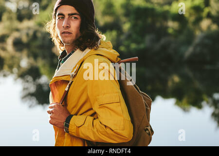 Seitenansicht eines Reisenden tragen Rucksack stand neben einem See entfernt. Nahaufnahme von einem jungen Mann, der in der Nähe von einem See auf eine Erforschung Reise. Stockfoto