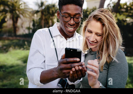 Lächelnd gemischtrassiges Paar stehen zusammen und Betrachten der Bilder auf dem Smartphone. Junger Mann und Frau an die Fotos auf Handy suchen Stockfoto