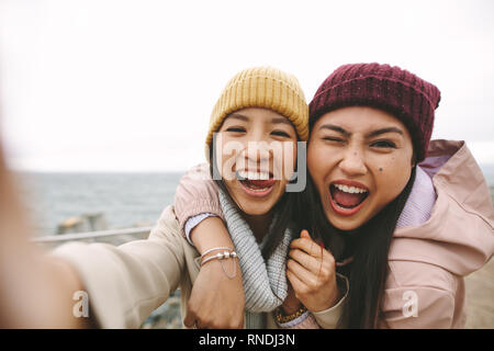 Freundliche asiatische Frauen im Winter tragen Spaß, die Gesichter in der Nähe von dem Meer. Lächelnde Mädchen Freunde stehen einander im Freien halten an einer Wint Stockfoto