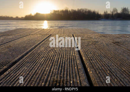 Fiery orange Sonnenuntergang auf einem ruhigen See mit Silhouettiert winter bäume niedrigen Winkel über die hölzerne strukturierte Platten einer Anlegestelle gesehen reflektiert Stockfoto