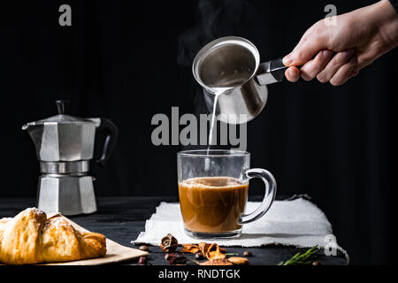 Gießen dampfende Milch in eine Tasse Kaffee. Hinzufügen von heißer Milch in den Espresso in italienischen Moka gebraut, Low-key-shot Stockfoto